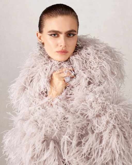 Close-up of a fashion model wearing a feathered coat, statement earrings, and a cocktail ring against a light neutral background.