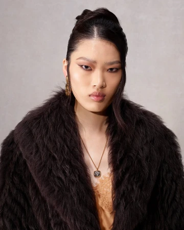 Model wearing dark brown fur collar coat and gold pendant necklace against a textured neutral background