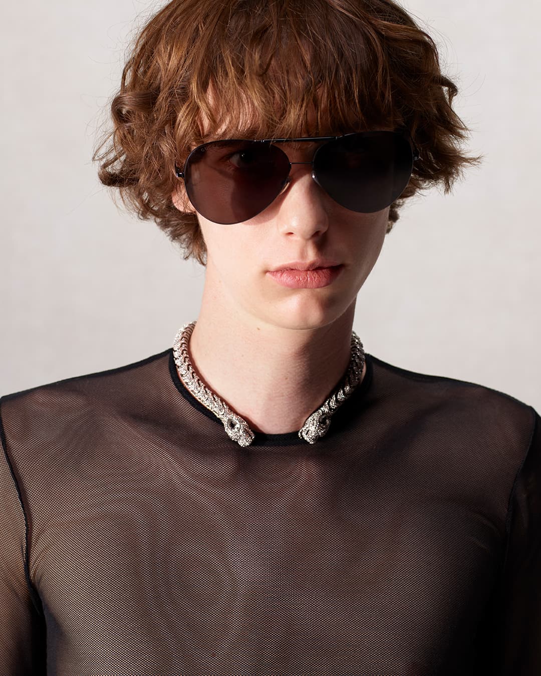 Male model wearing a black mesh top and a silver snake necklace, posing against a neutral backdrop, showcasing modern fashion.