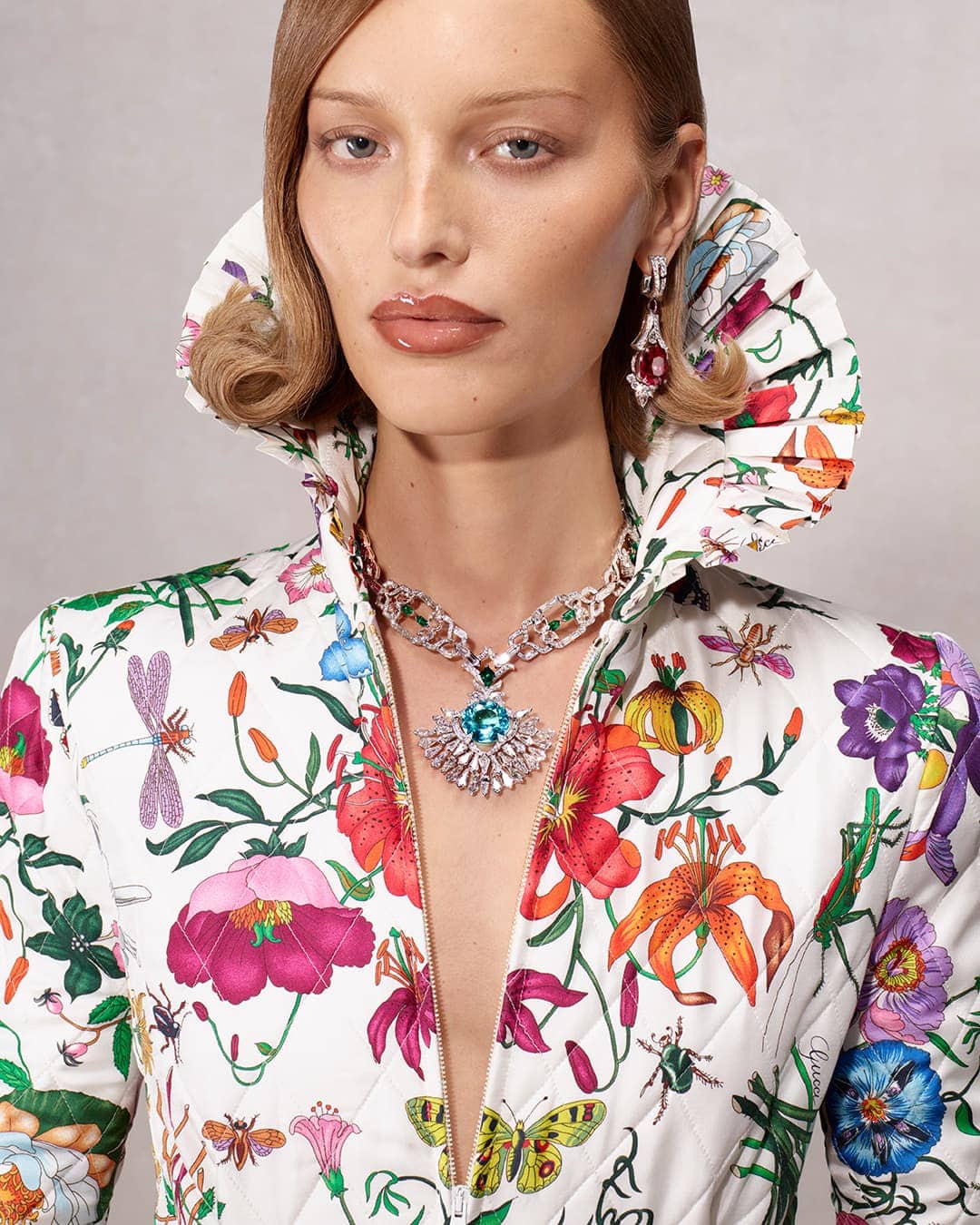 Close-up portrait of a woman wearing a white floral patterned jacket with a high ruffled collar, diamond necklace, and gemstone earrings.