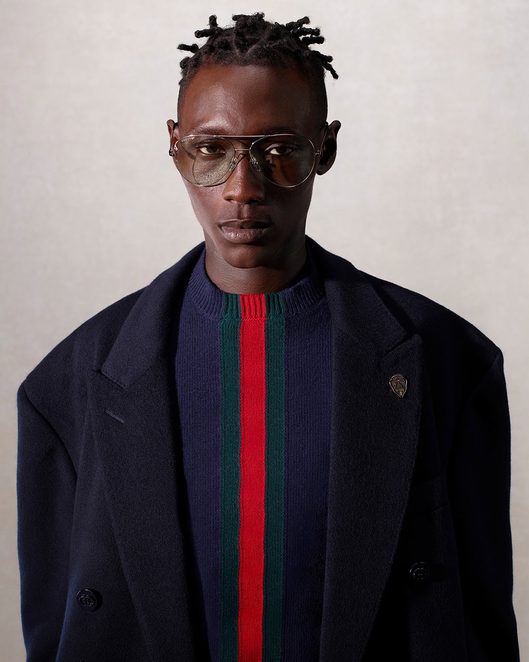 Portrait of a dark-skinned male model wearing aviator glasses, a striped sweater, and a dark coat in a neutral studio setting.