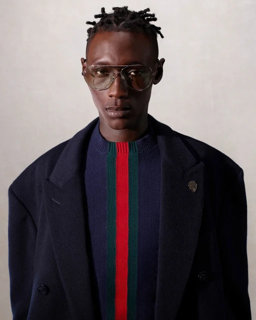 Portrait of a dark-skinned male model wearing aviator glasses, a striped sweater, and a dark coat in a neutral studio setting.