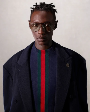 Portrait of a dark-skinned male model wearing aviator glasses, a striped sweater, and a dark coat in a neutral studio setting.