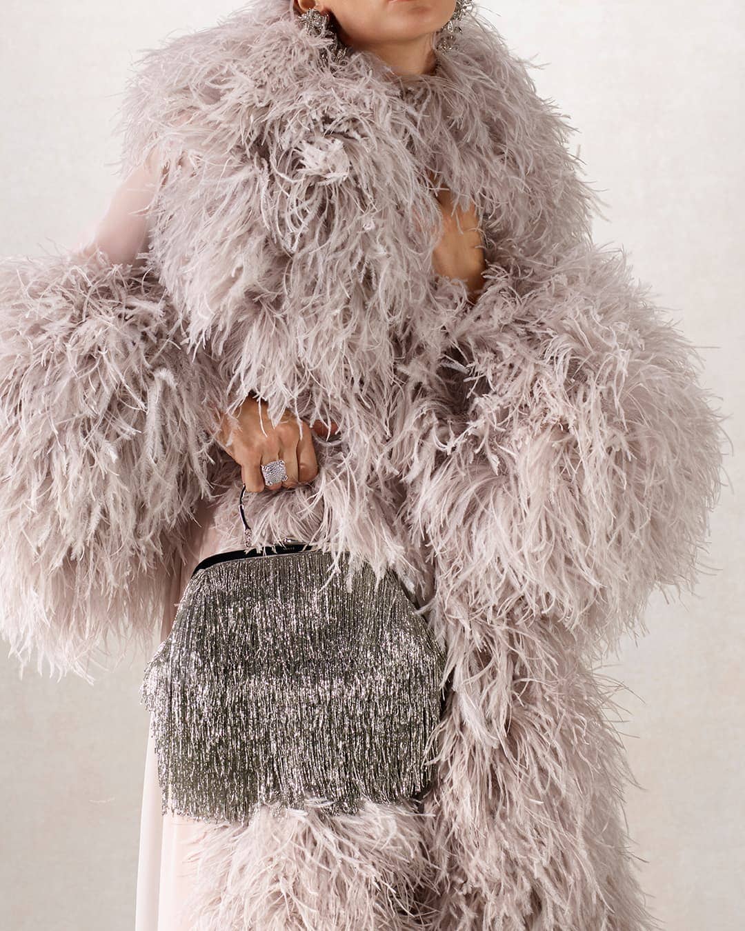 Elegant adult female wearing a feathered coat holds a fringed handbag in a neutral studio setting. Fashion and style are on display.