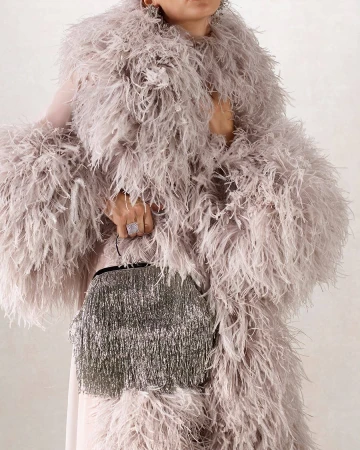 Elegant adult female wearing a feathered coat holds a fringed handbag in a neutral studio setting. Fashion and style are on display.