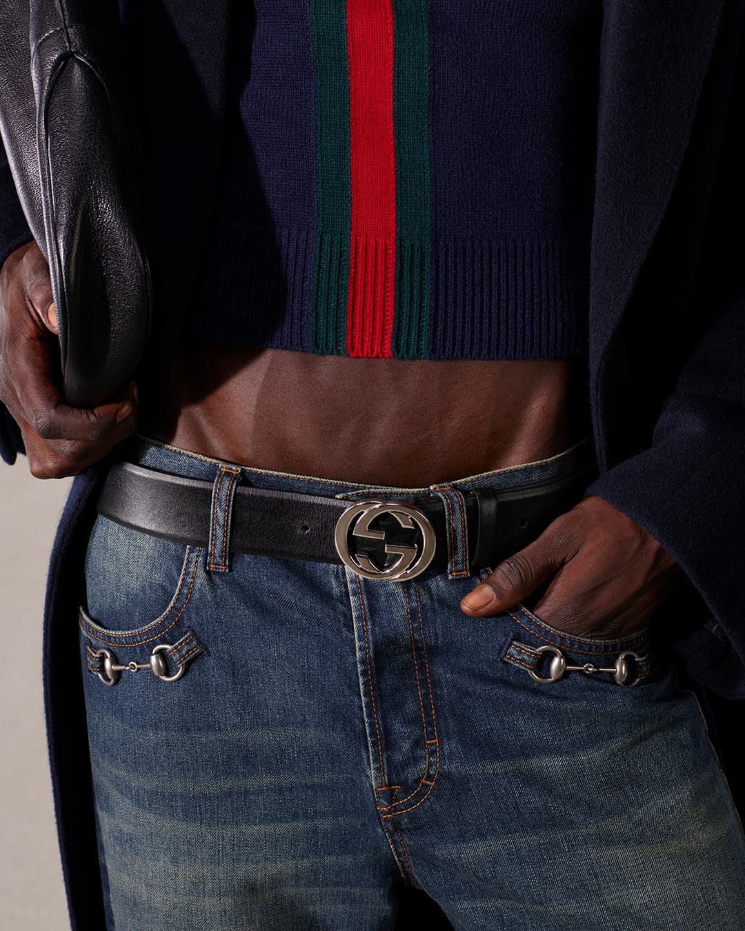 Close-up of a man wearing blue jeans and a Gucci belt with a silver buckle, also features a cropped top and a black coat.