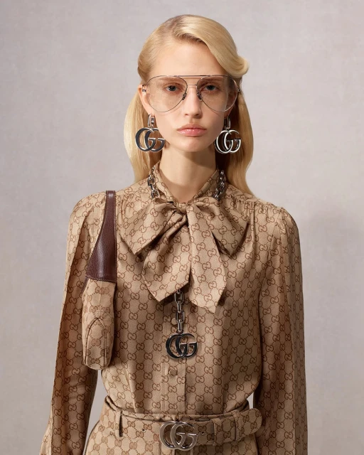 A blonde female model showcases a beige Gucci-patterned blouse with a bow, skirt, and matching accessories in a studio shot.