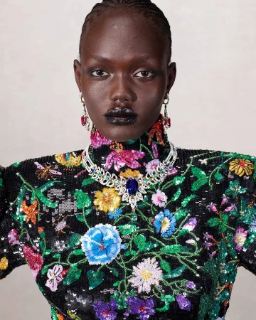 Close-up studio shot of an adult female model with dark skin and black lips wearing a high-necked dress with colorful floral embroidery.