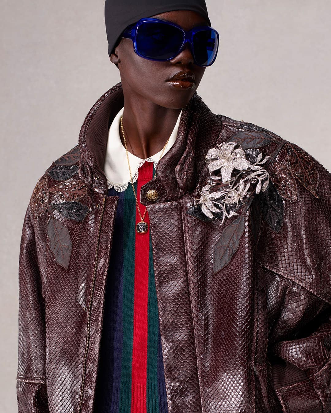 Fashion shot of an adult woman with dark skin wearing a snakeskin jacket, colorful sweater, black beanie, and sunglasses in a studio.