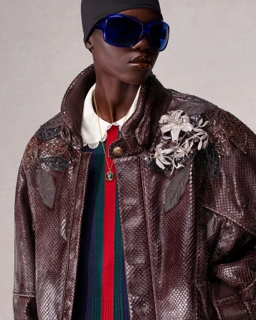 Fashion shot of an adult woman with dark skin wearing a snakeskin jacket, colorful sweater, black beanie, and sunglasses in a studio.