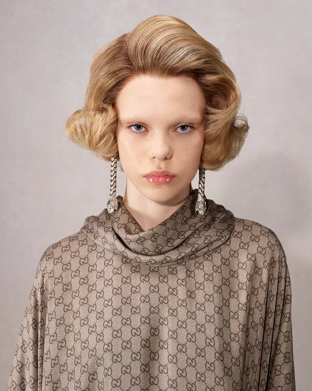 Blonde-haired model with flawless skin and statement earrings showcases a designer gown featuring a repeating logo pattern in a portrait.