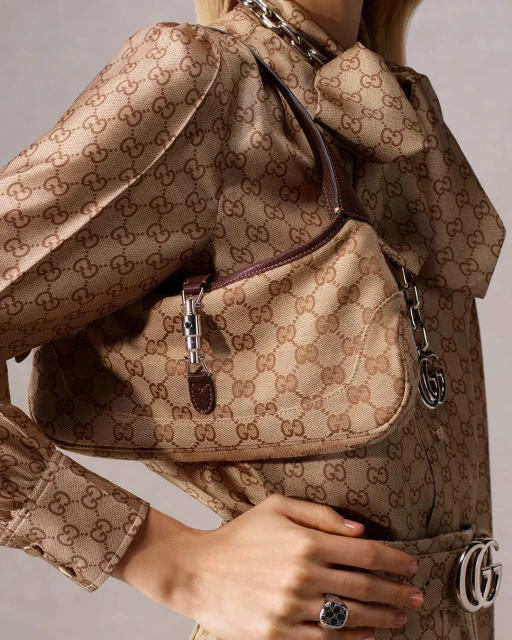 A Gucci Jackie bag is draped over a person wearing a dress with the Gucci logo pattern. A silver chain and silver ring add shine.