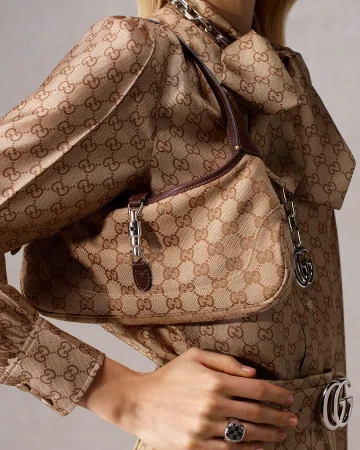 A Gucci Jackie bag is draped over a person wearing a dress with the Gucci logo pattern. A silver chain and silver ring add shine.