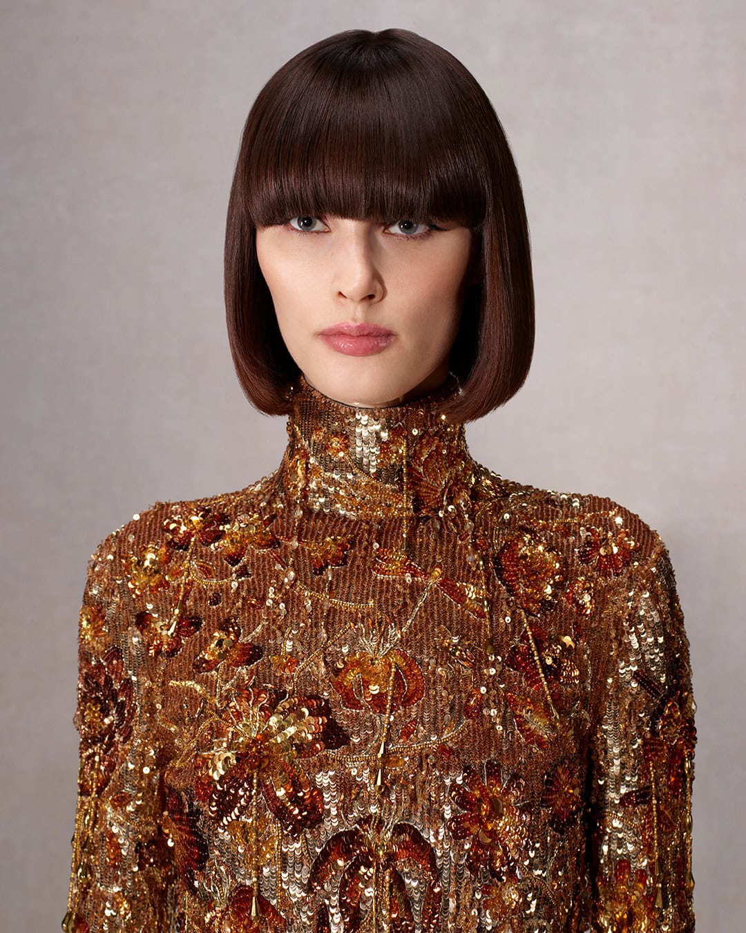 Woman with bob hairstyle and bangs wearing an embroidered high-neck dress in a studio with neutral backdrop, posing in portrait