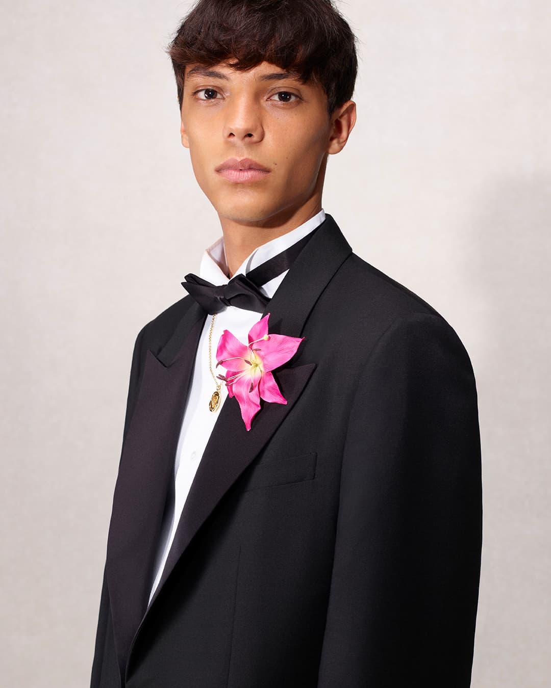 Young adult male in a black tuxedo and bow tie poses with a pink flower boutonniere in a studio setting with neutral lighting.