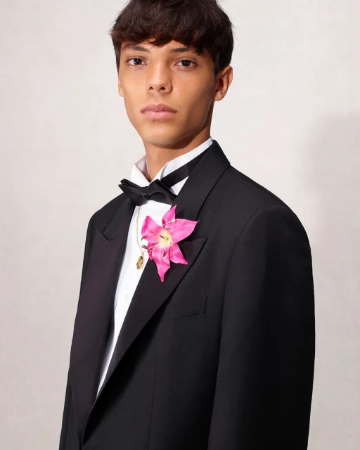 Young adult male in a black tuxedo and bow tie poses with a pink flower boutonniere in a studio setting with neutral lighting.