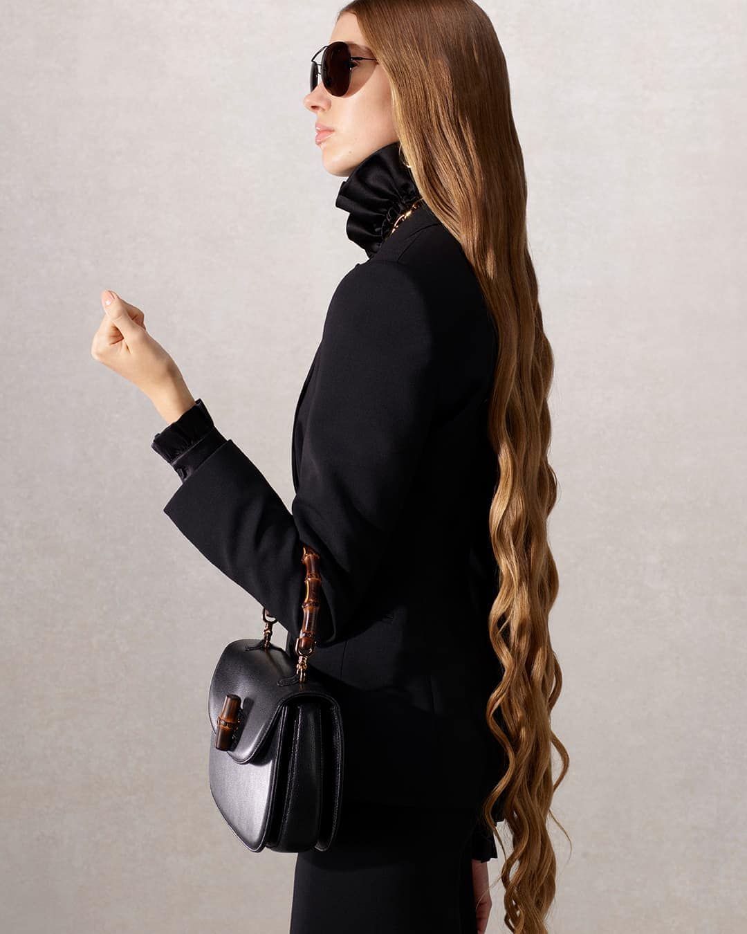 Profile view of an adult woman with long blonde hair wearing black suit and sunglasses, carrying a black leather purse with bamboo handle.