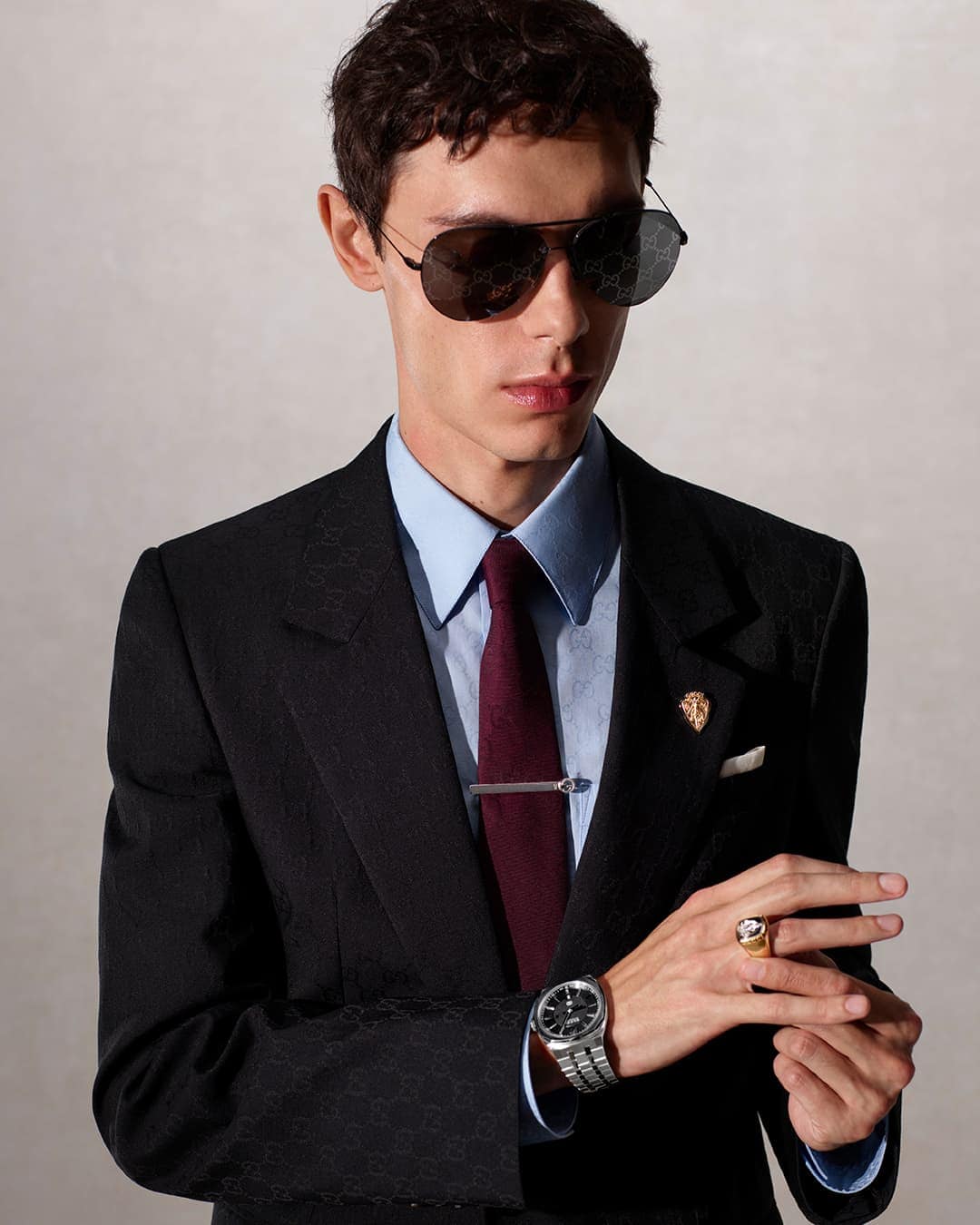 Young man in a black suit and sunglasses poses in a studio, wearing a light blue dress shirt and dark red tie, with a watch and ring.