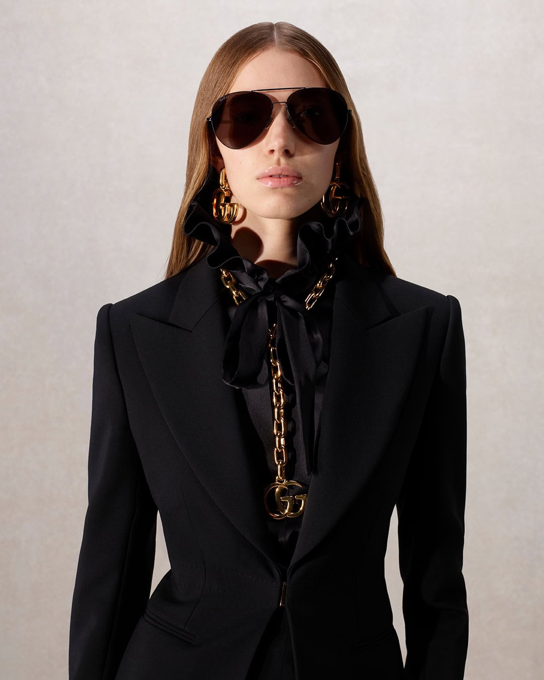 A woman wearing sunglasses, a black blazer, statement earrings, and a gold chain with a G pendant poses indoors against a neutral backdrop.