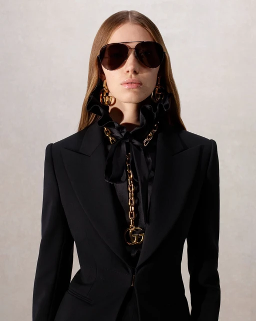 A woman wearing sunglasses, a black blazer, statement earrings, and a gold chain with a G pendant poses indoors against a neutral backdrop.