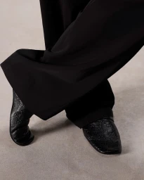 Close-up of black leather mules with embossed monogram pattern and wide trousers on a neutral floor.