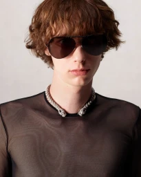 Model wearing dark aviator sunglasses and an ornate silver animal-head choker necklace against a plain grey backdrop.