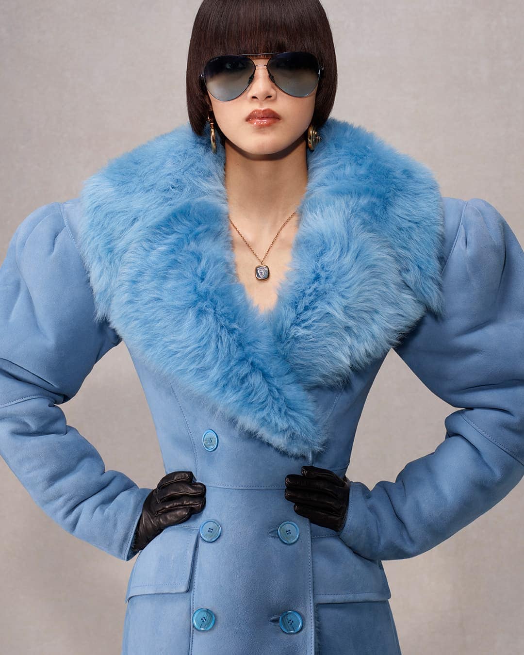 Model in blue coat with fur collar wearing sunglasses and black gloves against a grey backdrop