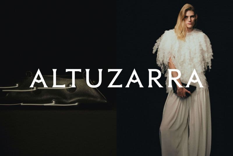Fashion shot of a model wearing a white fringed top and wide-leg pants, holding a black clutch, against a black backdrop next to a black bag.