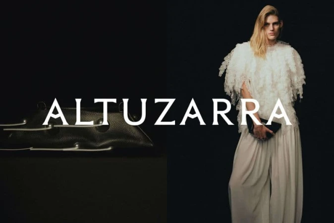 Fashion shot of a model wearing a white fringed top and wide-leg pants, holding a black clutch, against a black backdrop next to a black bag.