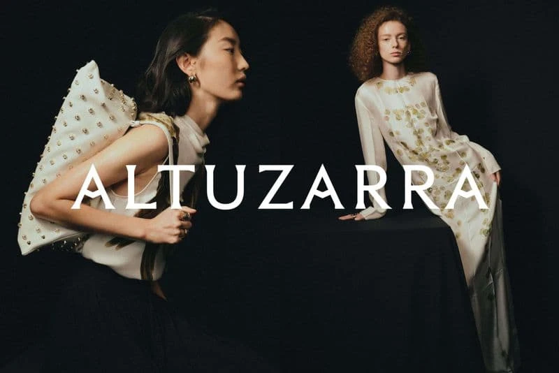 Two fashion models showcase Altuzarra designs against a dark backdrop, with one model wearing a studded bag and the other a patterned dress.