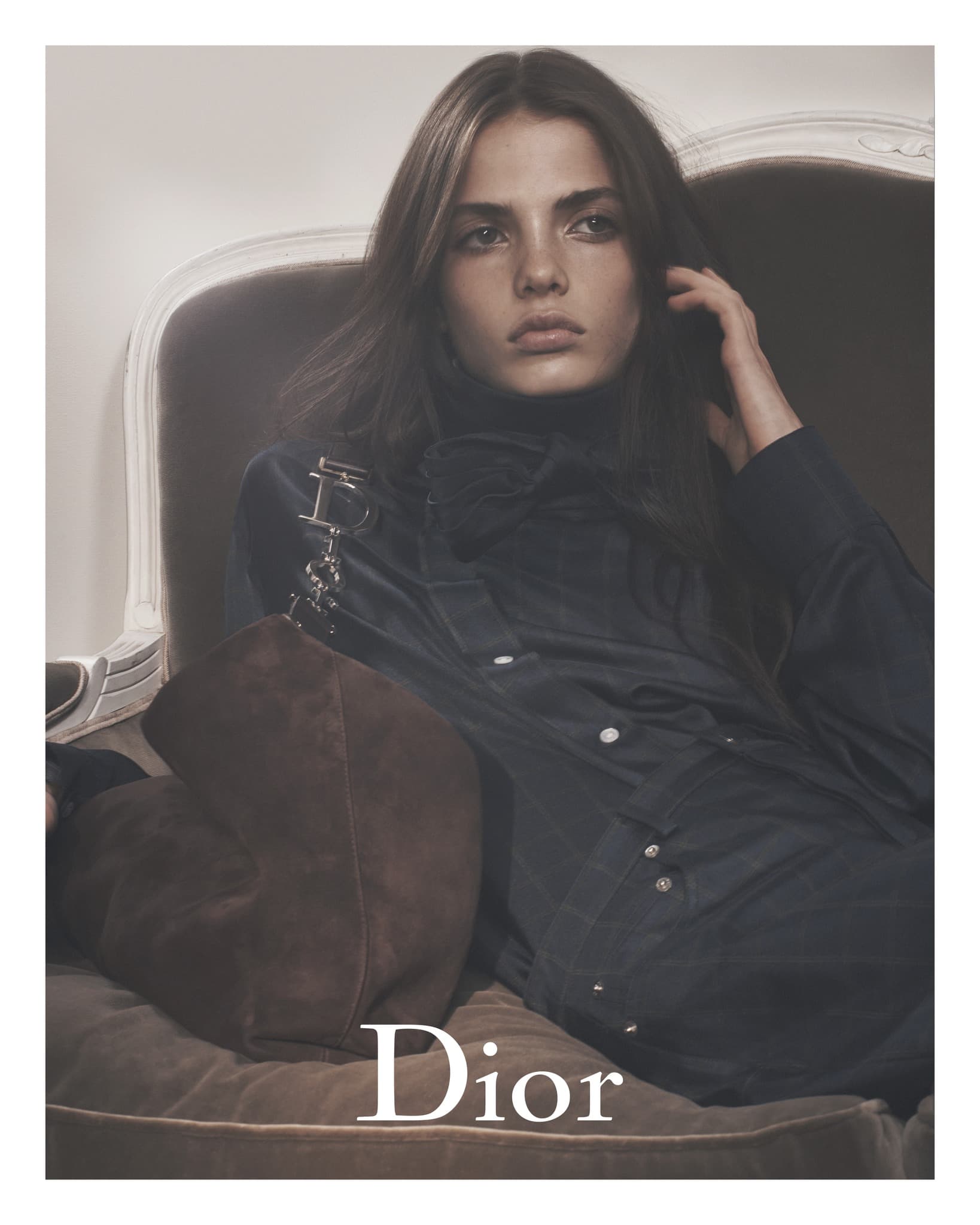 Fashion shot of a brunette woman in a black top and gray pants, sitting on a sofa with a brown handbag.  Dior logo is visible.