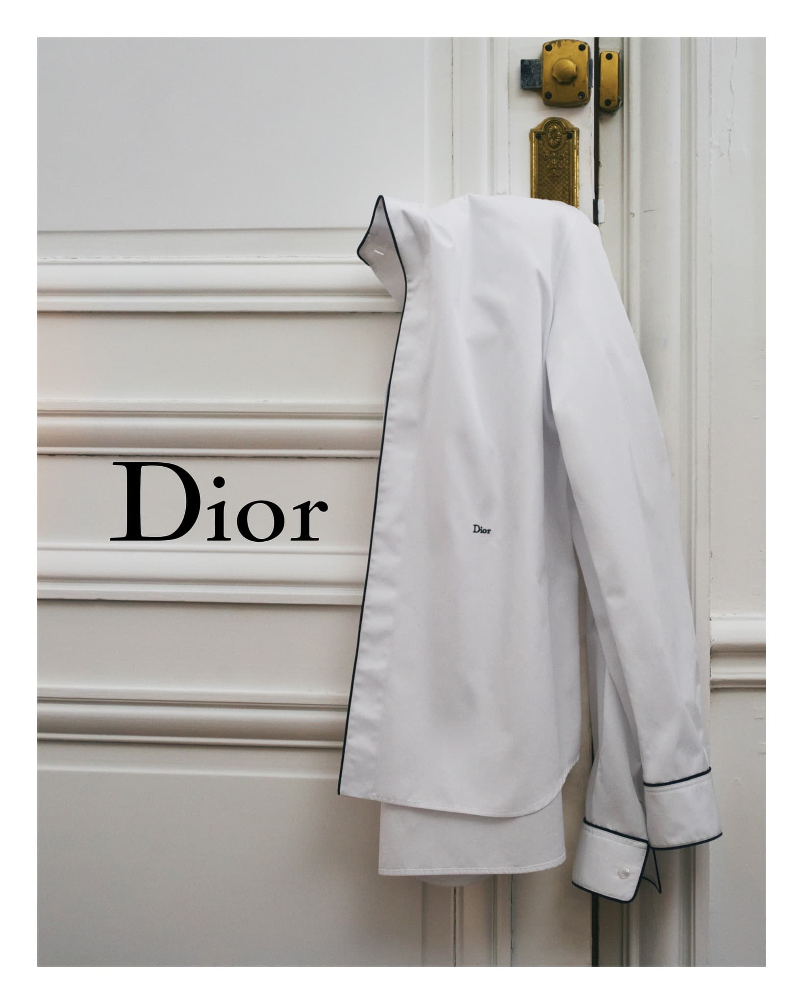 White collared shirt with dark trim hanging on white panelled door with bronze hardware and black Dior logo, clean presentation, fashion shoot.