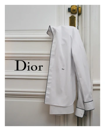 White collared shirt with dark trim hanging on white panelled door with bronze hardware and black Dior logo, clean presentation, fashion shoot.