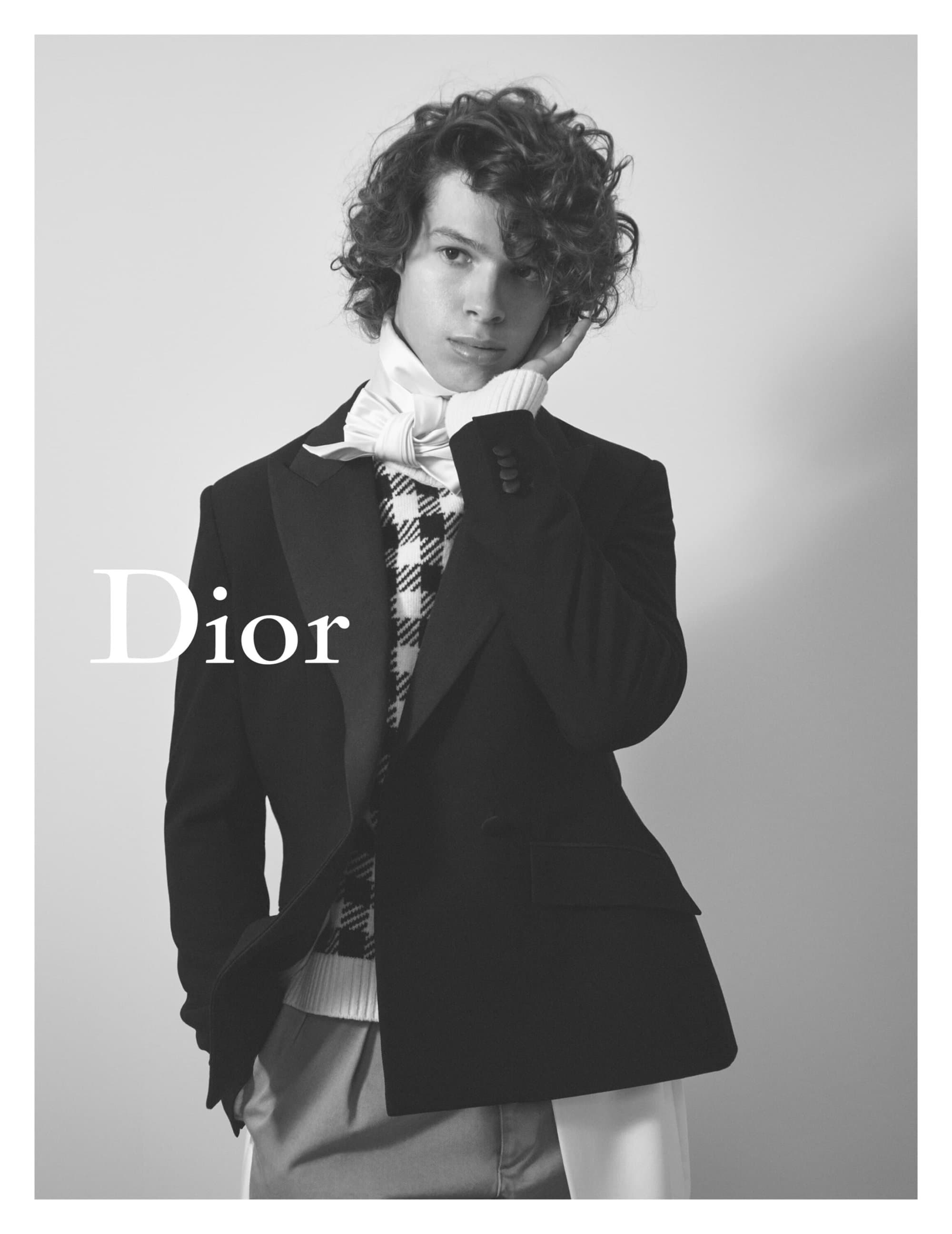 Black-and-white studio shot of a curly-haired young adult male model in a blazer, sweater vest, collared shirt, and trousers, with the Dior logo.