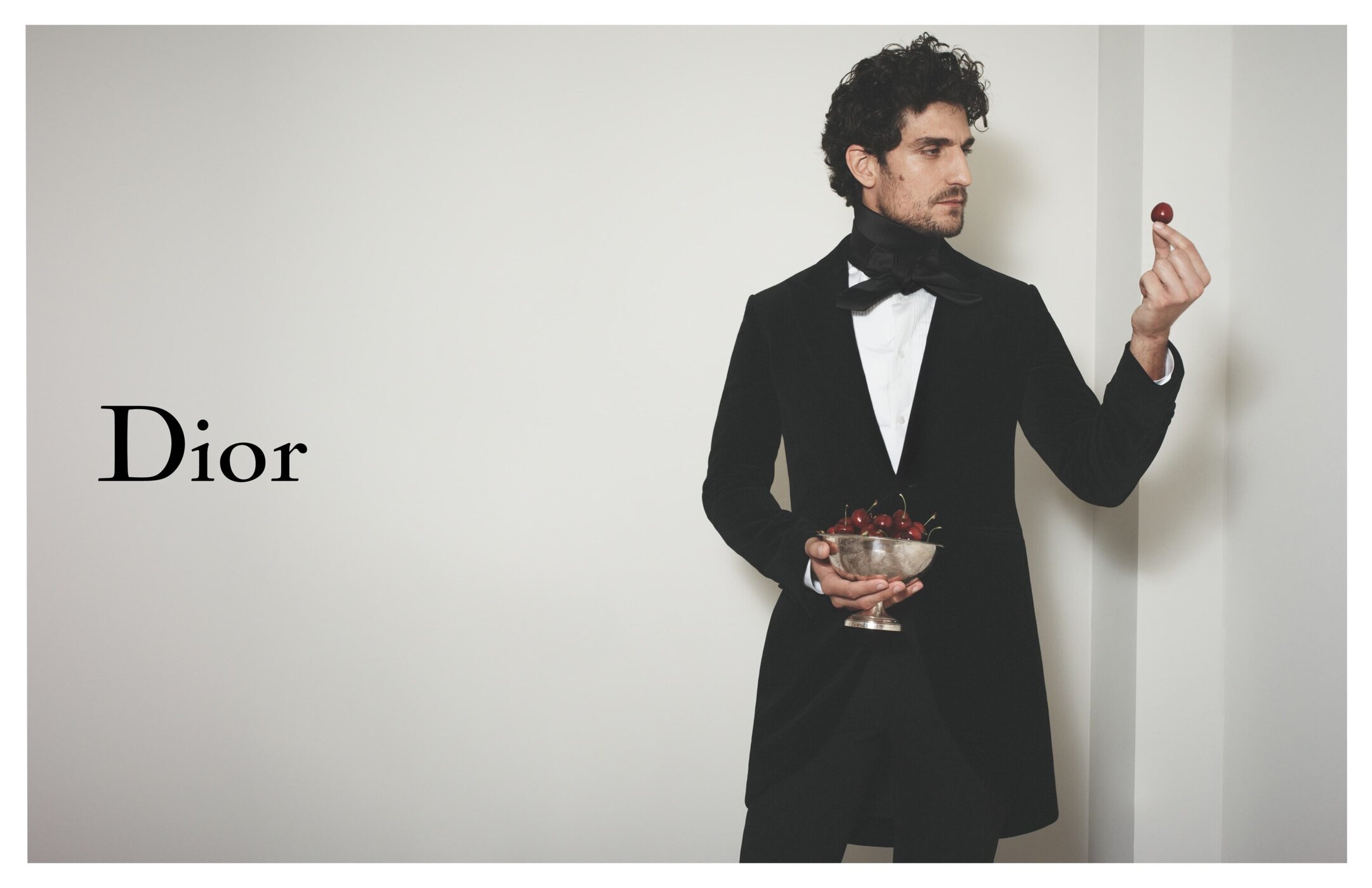 Man in black suit holds bowl of cherries indoors against a plain background for Dior, fashion-related content with neutral studio lighting.