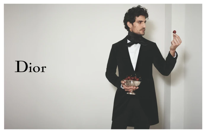 Man in black suit holds bowl of cherries indoors against a plain background for Dior, fashion-related content with neutral studio lighting.