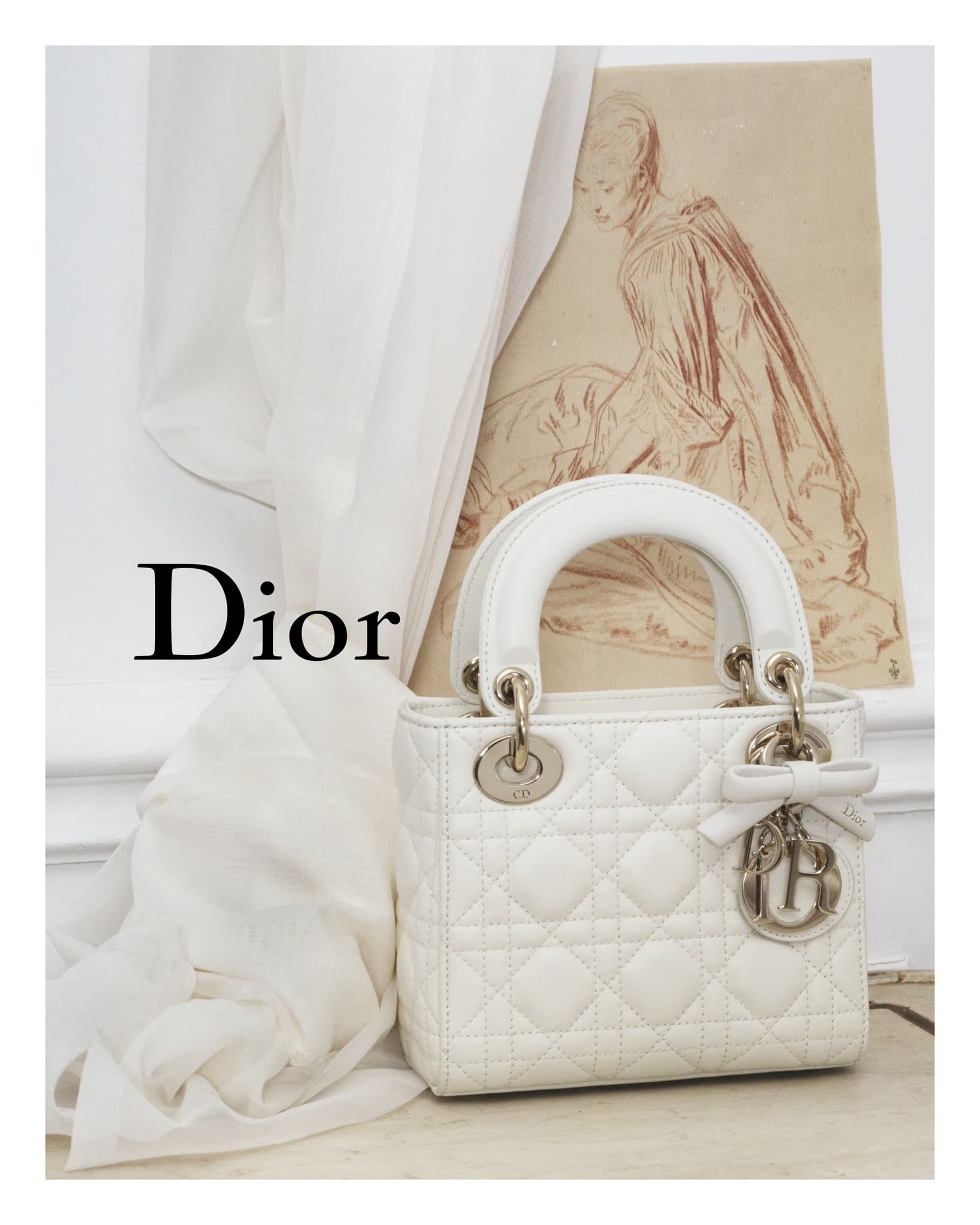 White Dior Lady Dior handbag with silver hardware, bow detail, set against a white curtain and art piece.