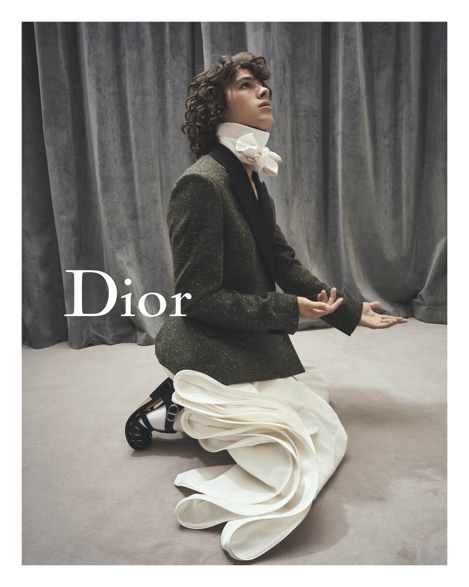 A person in a suit kneeling and looking upwards, with a large white decorative bow at the neck, Dior logo visible, and soft lighting.
