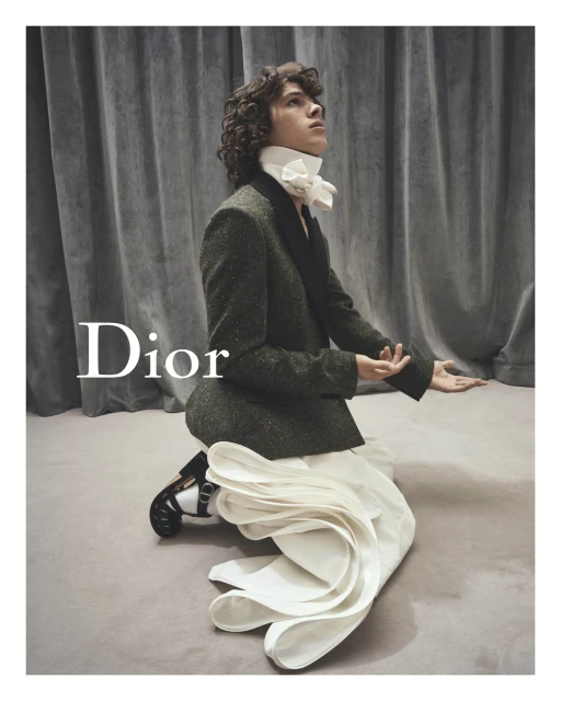 A person in a suit kneeling and looking upwards, with a large white decorative bow at the neck, Dior logo visible, and soft lighting.