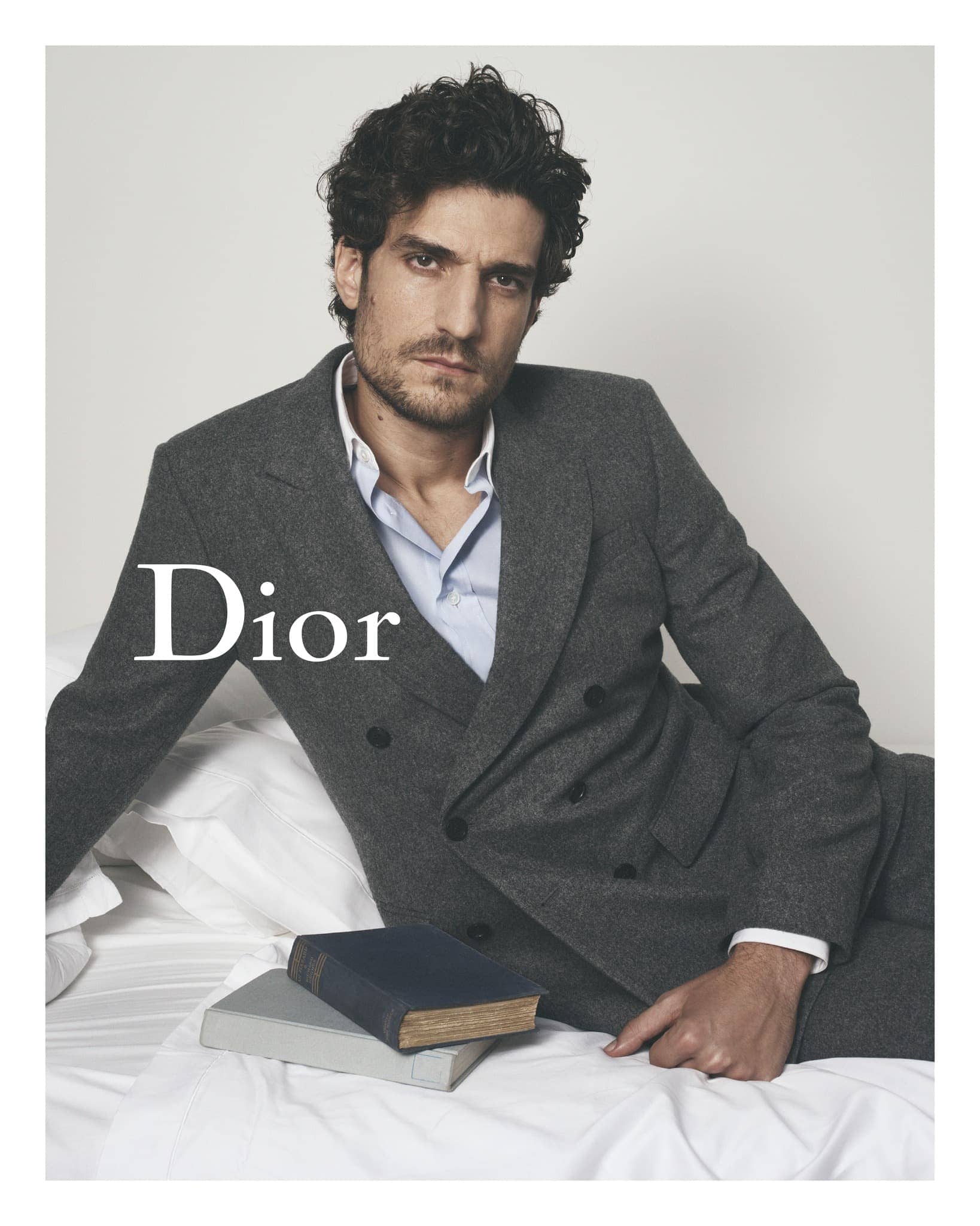 Adult male in grey suit and blue shirt lies on a white bed next to two books. Fashion editorial-style image for Dior ad campaign.