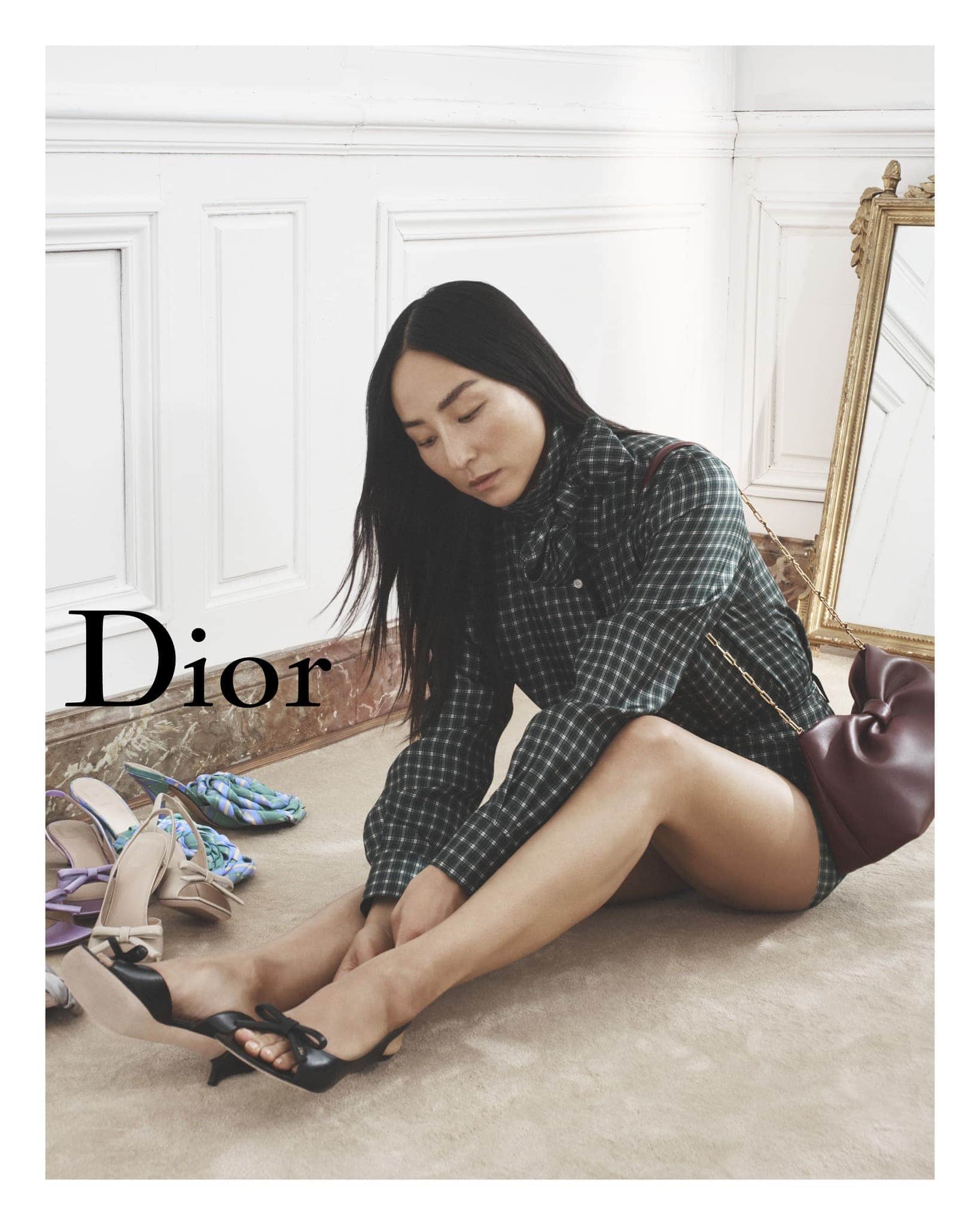 Asian woman in a plaid top sits on a carpet surrounded by assorted high-heeled shoes, a shoulder bag and a gilded mirror behind.