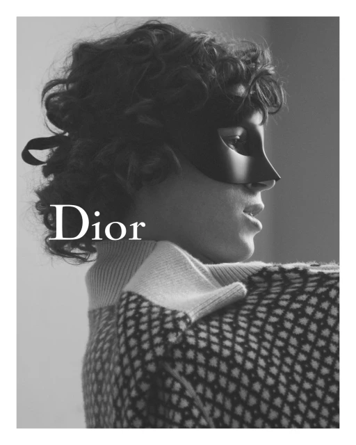 Black and white portrait of a woman with curly hair wearing a masquerade mask, diamond patterned sweater, and the brand logo, Dior.
