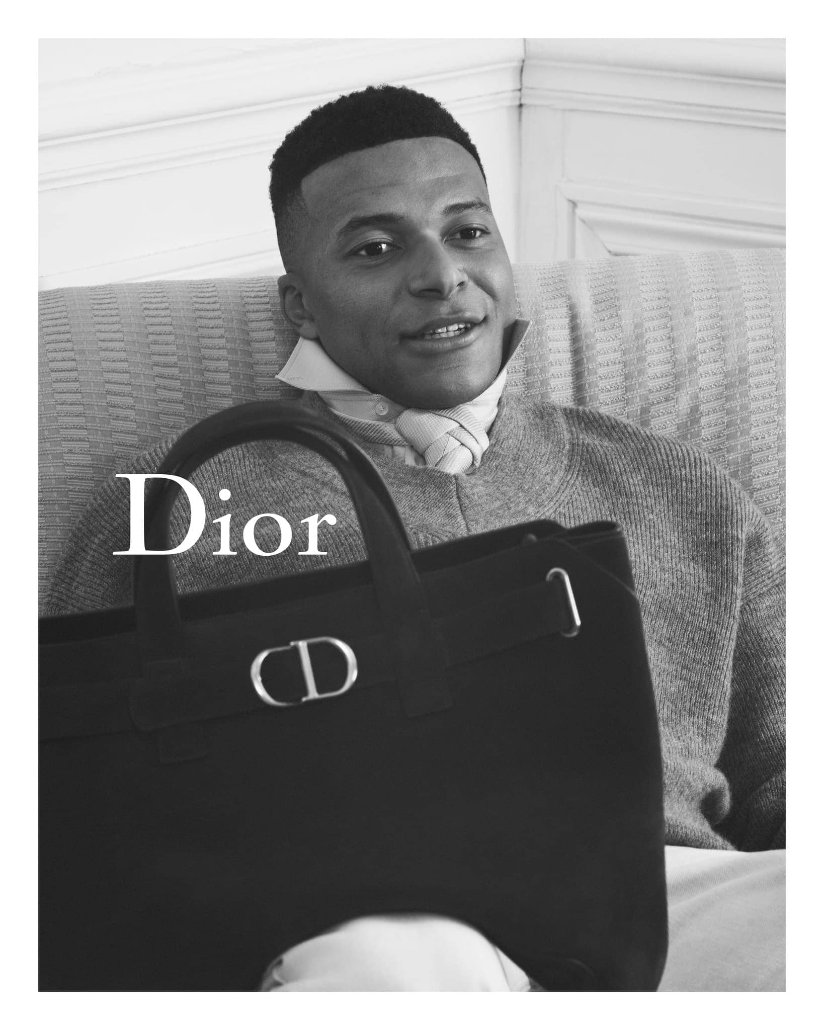 Adult male poses indoors in a medium shot, wearing a grey sweater, holding a black Dior handbag. The setting is minimalist.