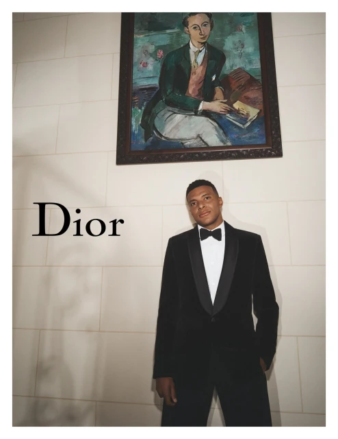 Man in black tuxedo stands under portrait in an indoor setting, fashion brand advertisement, neutral color palette.