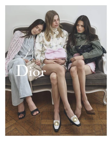 Three fair-skinned women seated on a grey couch wearing fashionable clothes and shoes, indoor, medium shot. Dior logo is visible.