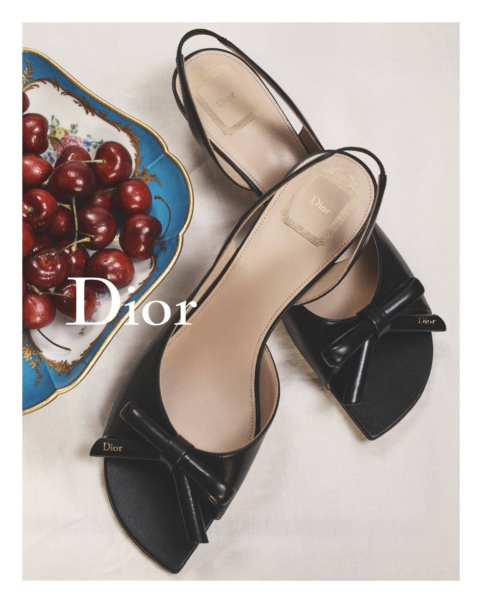 High-angle shot of two black patent leather slingback shoes with bow details and a tray of cherries, showcasing luxury fashion footwear.