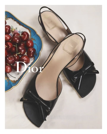 High-angle shot of two black patent leather slingback shoes with bow details and a tray of cherries, showcasing luxury fashion footwear.