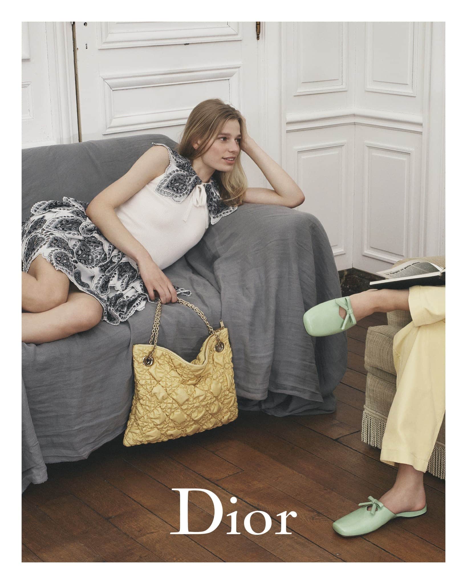 Woman in a sleeveless white top and paisley skirt lounges on a gray couch with a yellow quilted bag. Another person wears green mules.