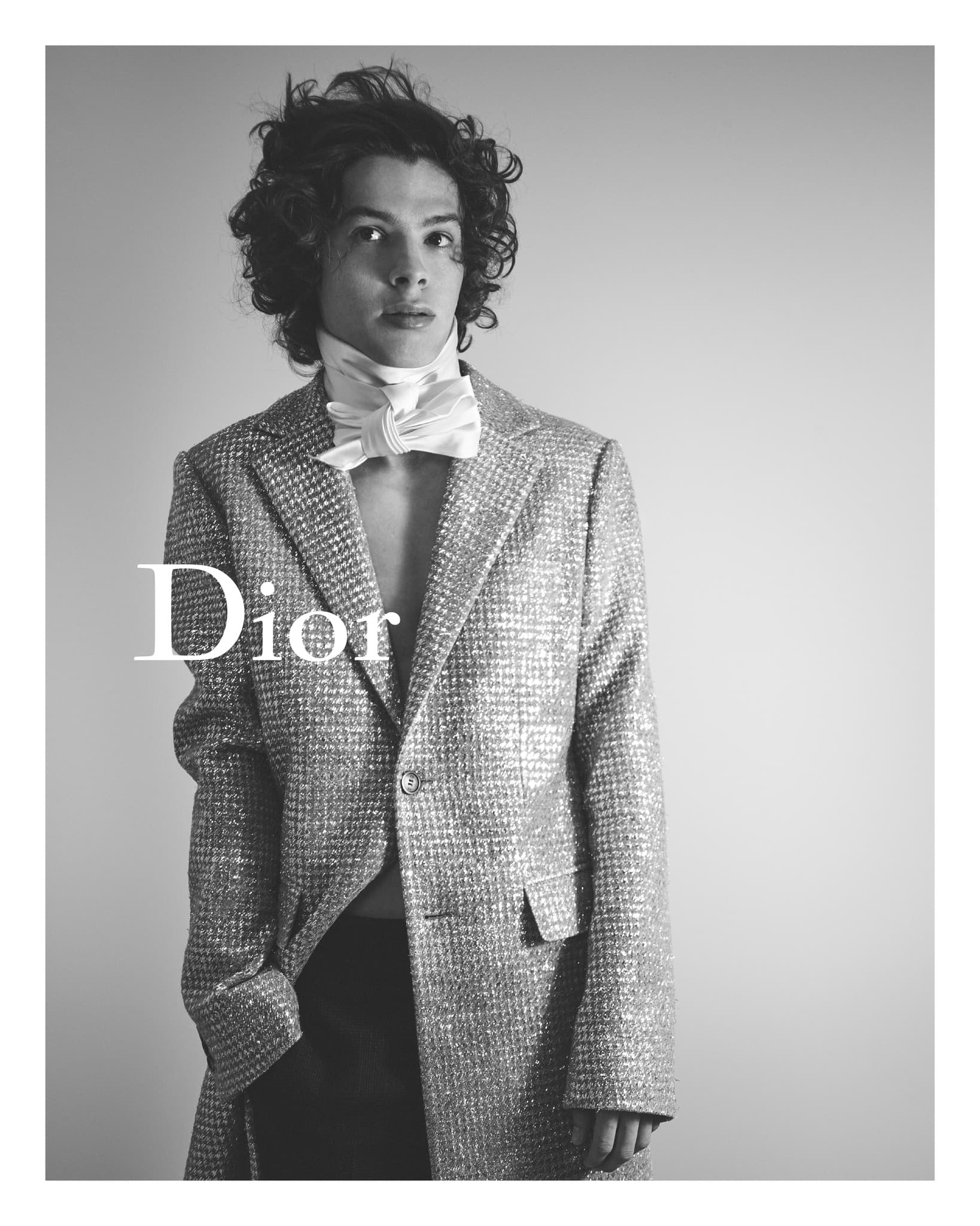 Black and white portrait of a young man with curly hair wearing a textured jacket and white bowtie, posing in front of a plain backdrop.