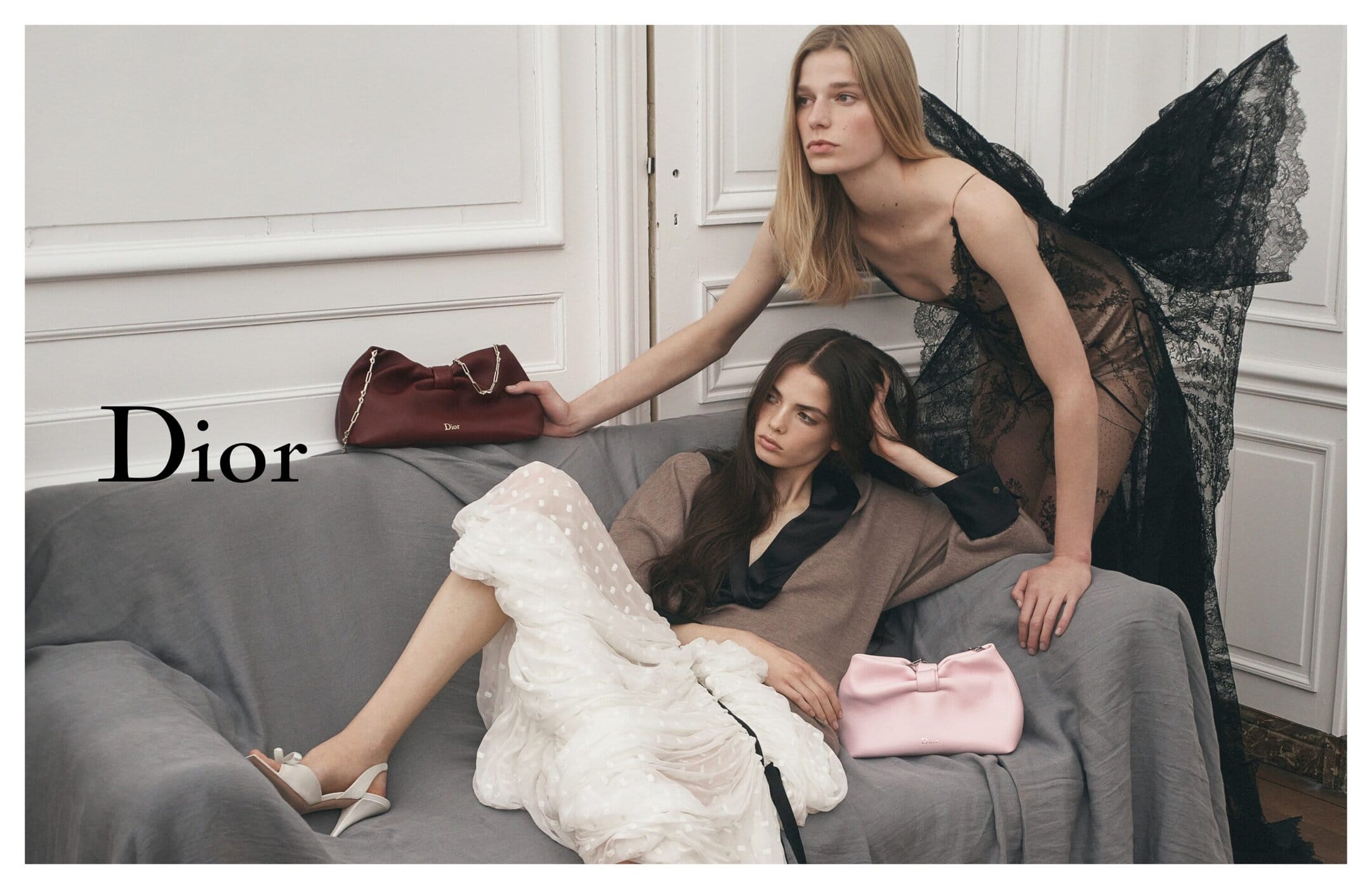 Two fashion models pose on a grey couch, wearing a brown sweater and a black lace dress. Two handbags are placed on the couch.